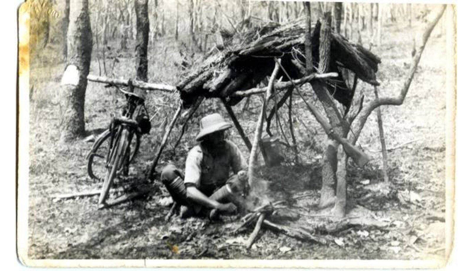 Czarno-białe zdjęcie przedstawia mężczyznę, który rozpala ognisko. Siedzi obok prowizorycznego szałasu, o który oparty jest rower.