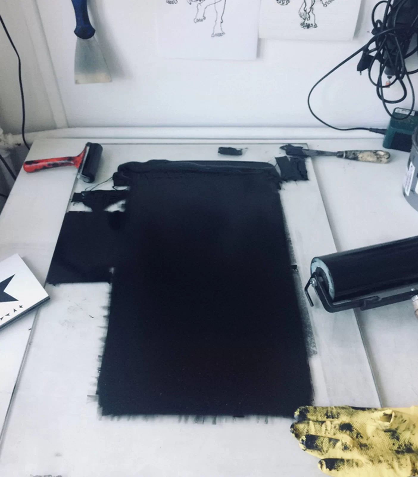 Ilustracja przedstawia leżący na stole arkusz linoleum zamalowany na czarno wałkiem. Obok leżą narzędzia pracy: dwa różnej wielkości wałki do nakładania farby oraz szpachelka. Druga szpachla wisi na ścianie. Po przeciwnej stronie znajduje się zwój kabli. W dolnym, prawym rogu widoczny jest fragment dłoni w żółtej, gumowej rękawicy ubrudzonej drukarską, czarną farbą.