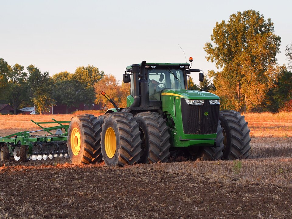 Zdjęcie przedstawia ciągnik rolniczy. Jest on zielony o wysuniętej przedniej części. Na dachu ma lampkę kontrolną. Z tyłu ciągnie urządzenie do orania gleby. Traktor znajduje się na błotnistej ziemi porośniętej lekką trawą. W tle znajdują się zabudowania rolnicze oraz wysokie drzewa.