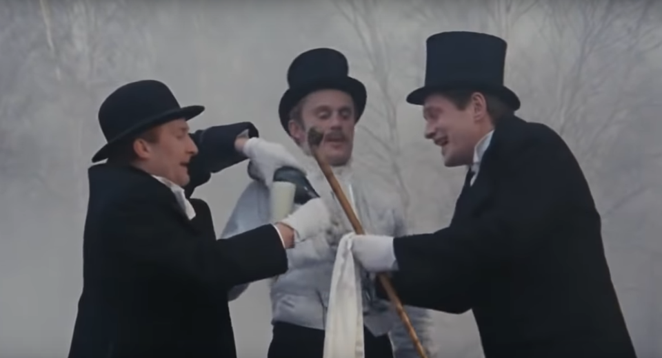 Kadr z filmu Ziemia obiecana. Trzech mężczyzn ubranych w odświętne stroje: ciemne płaszcze, białe rękawiczki, cylindry i meloniki. Stoją na zewnątrz, w zamglonej przestrzeni. Jeden z nich nalewa napój do kieliszka, który trzyma drugi. Trzeci mężczyzna opiera się o laskę, na której zawieszona jest biała tkanina. W tle widoczne są nagie gałęzie drzew. Mężczyźni są uśmiechnięci, zwróceni do siebie. Postacie ustawione są blisko siebie.