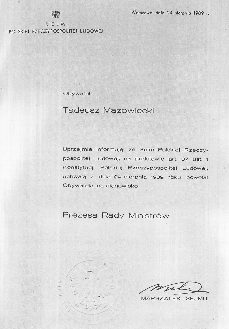 Zdjęcie przedstawia akt prawny. Jest to powołanie Tadeusza Mazowieckiego na stanowisko Prezesa Rady Ministrów. Dokument został odręcznie podpisany przez Marszałka Sejmu. Organem powołującym nowego premiera był Sejm Polskiej Rzeczpospolitej Ludowej. Powołanie na urząd Premiera miało miejsce dwudziestego czwartego sierpnia tysiąc dziewięćset osiemdziesiątego dziewiątego roku, w Warszawie. Treść dokumentu jest następująca: Obywatel Tadeusz Mazowiecki. Uprzejmie informuję, że Sejm Polskiej Rzeczpospolitej, na podstawie artykułu 37 ust. 1 Konstytucji Polskiej Rzeczypospolitej Ludowej, uchwałą z dnia 24 sierpnia 1989 roku powołał Obywatela na stanowisko Prezesa Rady Ministrów. Podpisał Marszałek Sejmu. 