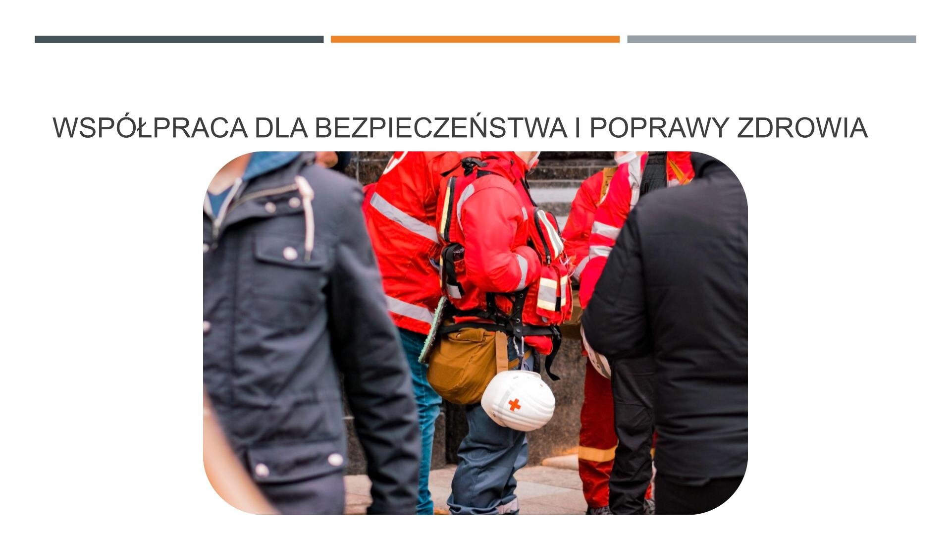 Biały slajd, na którym umieszczone zostało zdjęcie i tekst. Zdjęcie, znajdujące się w centralnej części slajdu, przedstawia dwóch mężczyzn ubranych w czerwone kurtki ratownicze. Na plecach mają czerwone kurtki, a do pasa przypięty biały kask z czerwonym krzyżem. Wokół przechodzą ludzie w ciemnych ubraniach. W górnej części slajdu czarny napis: „Współpraca dla bezpieczeństwa i poprawy zdrowia”.