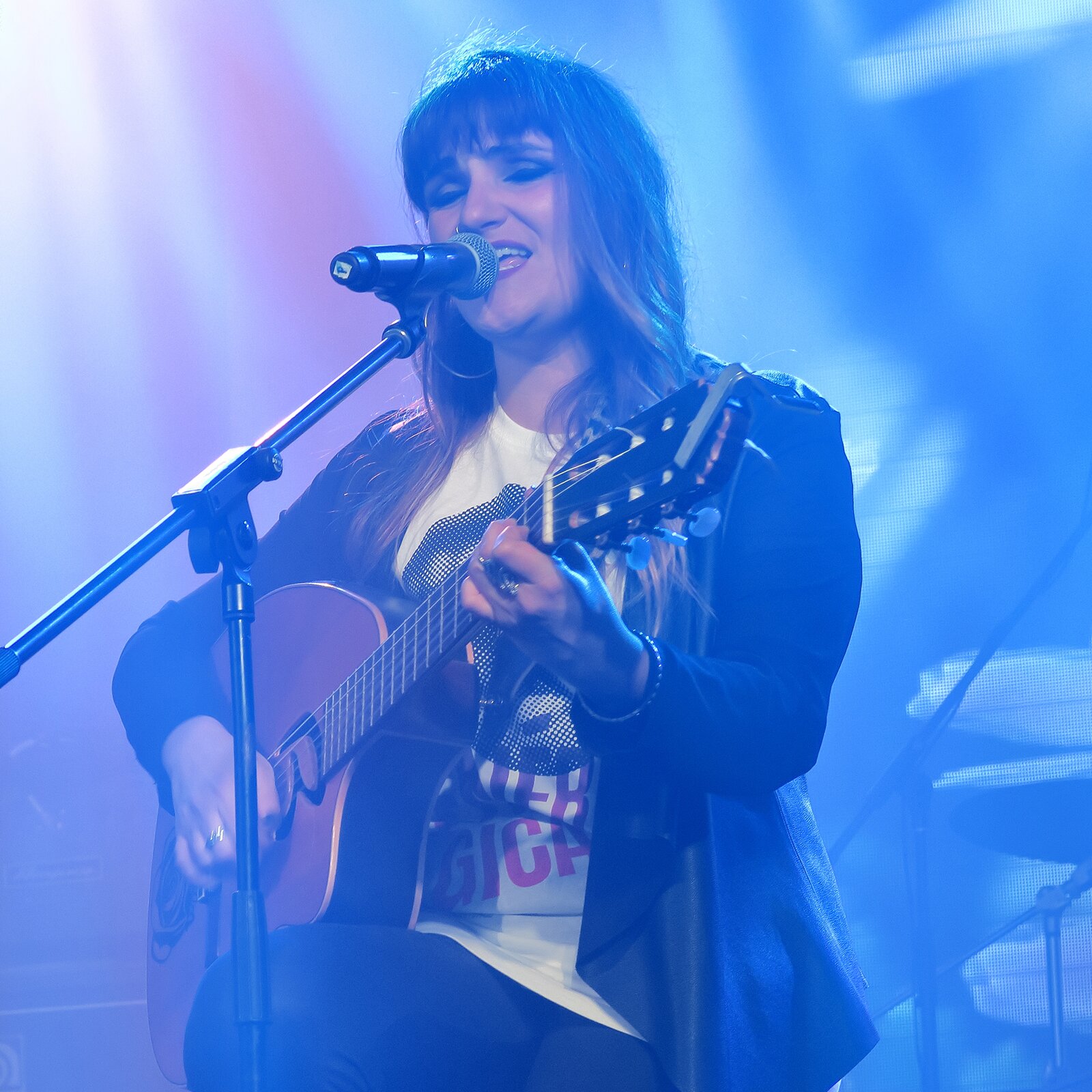 Zdjęcie przedstawia kobietę grającą na gitarze i śpiewającej do mikrofonu na scenie.