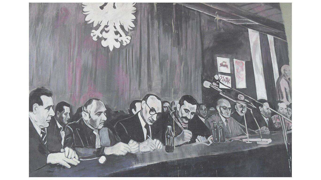 Fotografia czarno-biała przedstawiająca mural na wiadukcie w Gdańsku. Podpisanie umów sierpniowych w Gdańsku, w sali BHP w 1980 roku. Przy stole prezydialnym siedzą: w środku Lech Wałęsa z charakterystycznym, dużym długopisem, po jego lewej stronie Mieczysław Jagielski oraz inne postaci, które uczestniczyły w podpisaniu dokumentów.