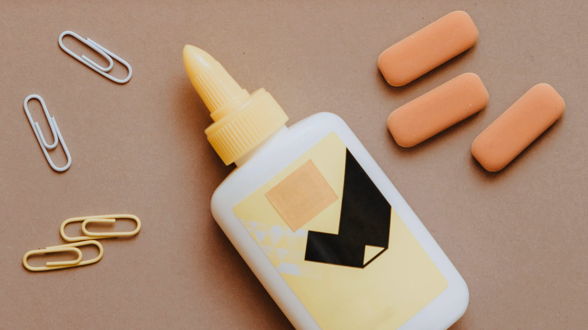Zdjęcie przedstawia mała butelkę z klejem. Obok leżą cztery spinacze i trzy płaskie gumki do ścierania.