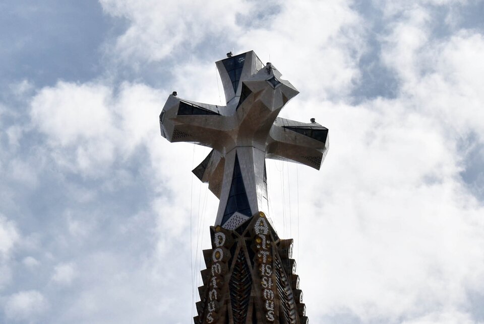 Ilustracja przedstawia Zwieńczenie Wieży Jezusa Chrystusa. Ukazuje górną część wieży bazyliki Sagrada Família, zakończoną dużym, lśniącym krzyżem o geometrycznym kształcie. Poniżej widać fragment masywnej, kamiennej konstrukcji wieży z drobnymi, precyzyjnymi zdobieniami. Tło stanowi jasne, lekko zachmurzone niebo.