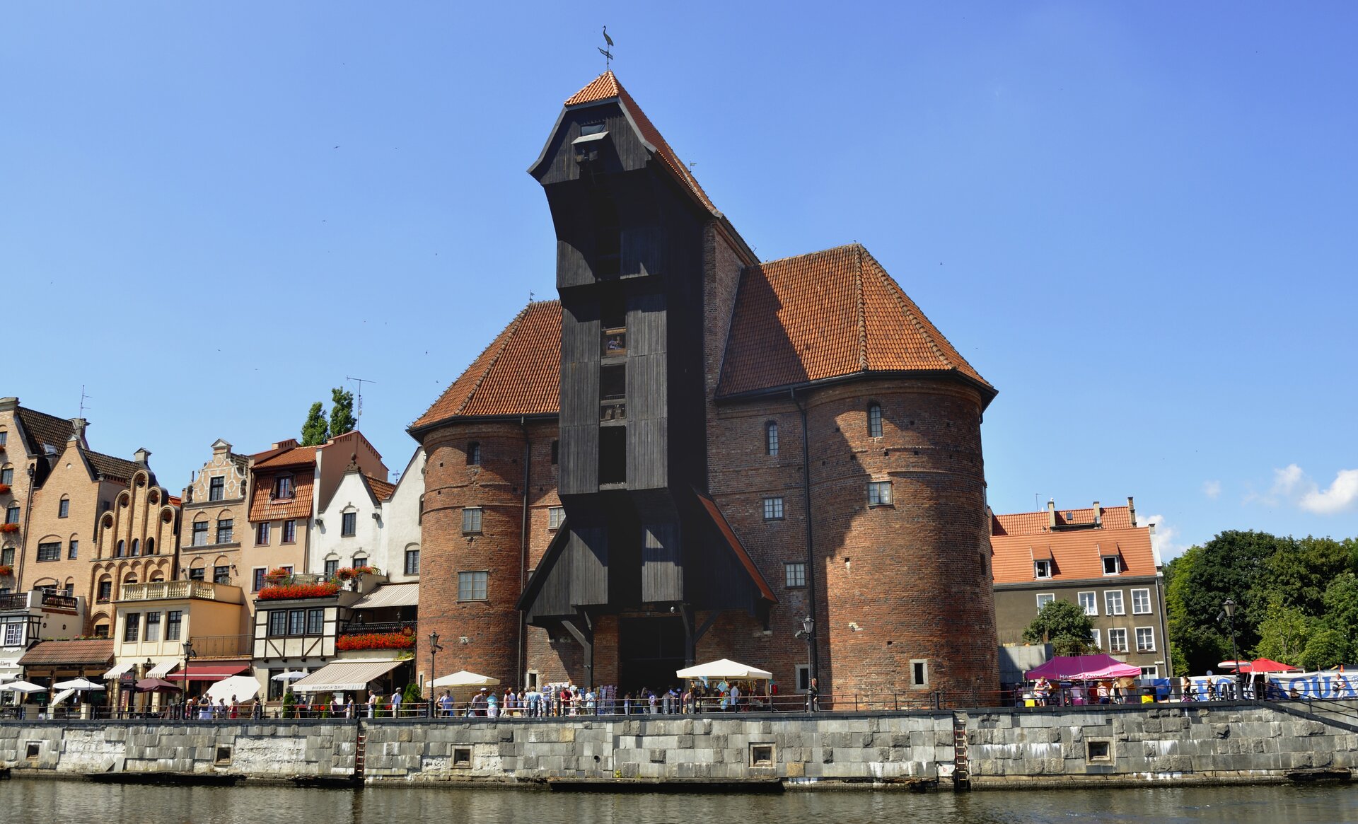 Fotografia barwna przedstawiająca gdański Żuraw od strony rzeki Motławy. Budynek ma około 30 m wysokości i 4 kondygnacje. Jego bryłę tworzą dwie masywne, okrągłe, ceglane baszty połączone mechanizmem dźwigowym. Jest on obudowany czarnym drewnem, wsparty na dwóch drewnianych filarach.  Color photograph of the Gdańsk Crane from the Motława River. The building is about 30 m high and has 4 floors. Its mass consists of two massive, round, brick towers joined by a crane mechanism. It is enclosed in black wood, supported on two wooden pillars.