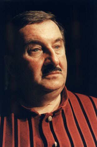 Na zdjęciu widać lewy profil pulchnej twarzy mężczyzny, który ma krótko przycięte, ciemne włosy, z przedziałkiem zaczesanym w prawą stronę. Nad ustami widać przycięty wąsik. Ubrany jest w czerwoną koszulę w pionowe czarne paski. Ma zaskoczony wyraz twarzy.