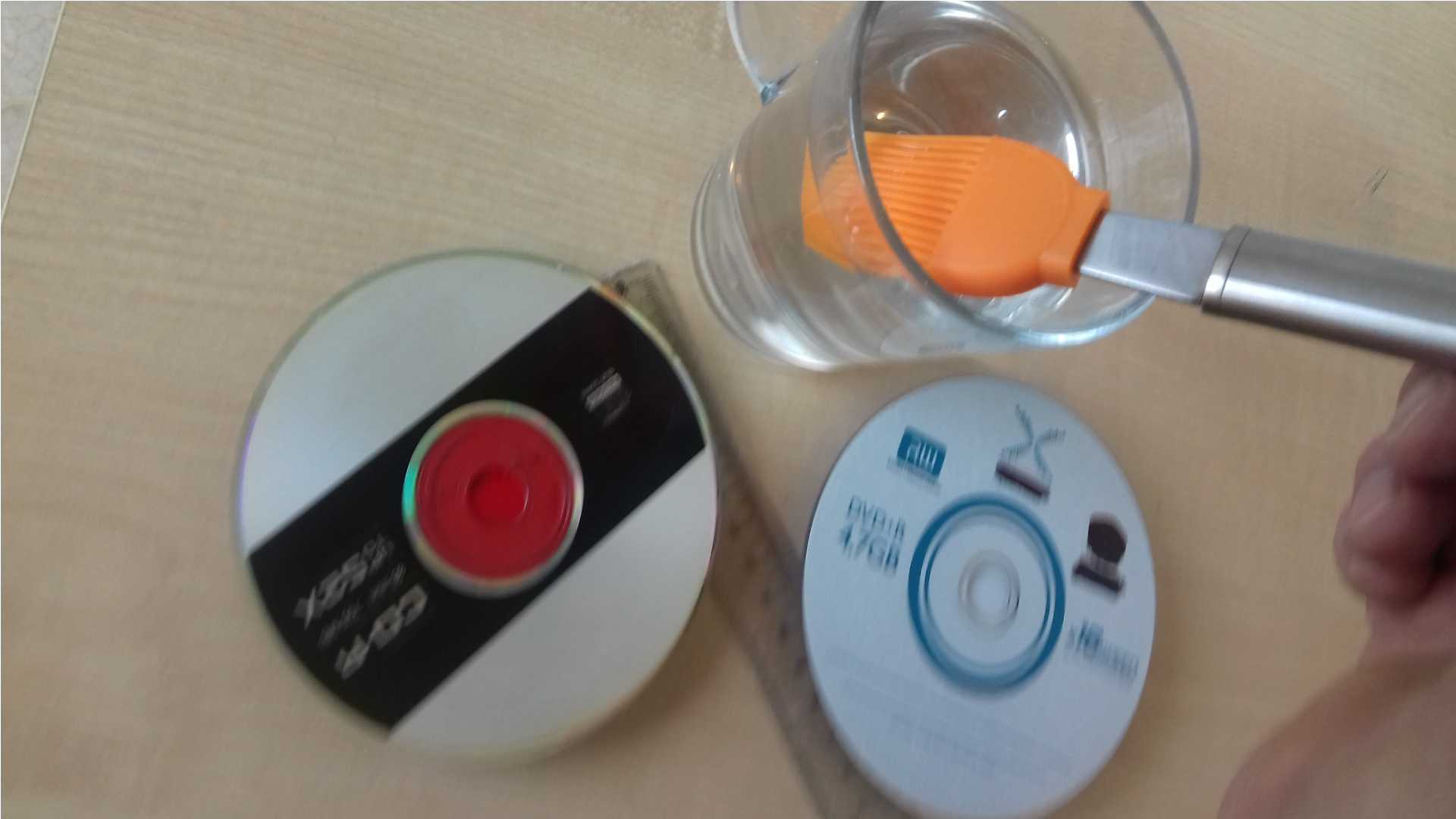 Zdjęcie przedstawia dwa dyski optyczne, linijkę oraz szklankę wody i pędzel w szklance wody.  