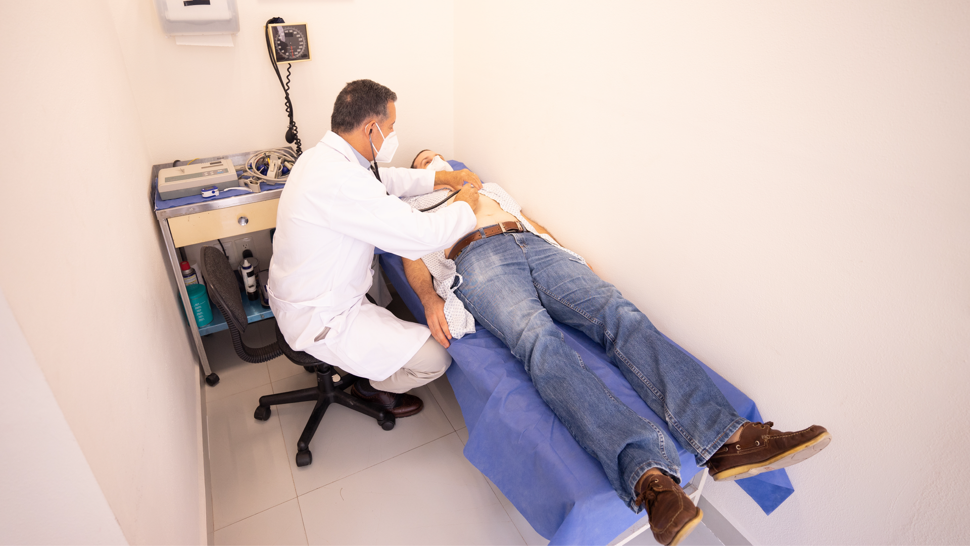 Zdjęcie przedstawia lekarza oraz młodego mężczyznę leżącego la leżance. Mężczyźni znajdują się w gabinecie lekarskim. Doktor bada pacjenta przy pomocy stetoskopu.