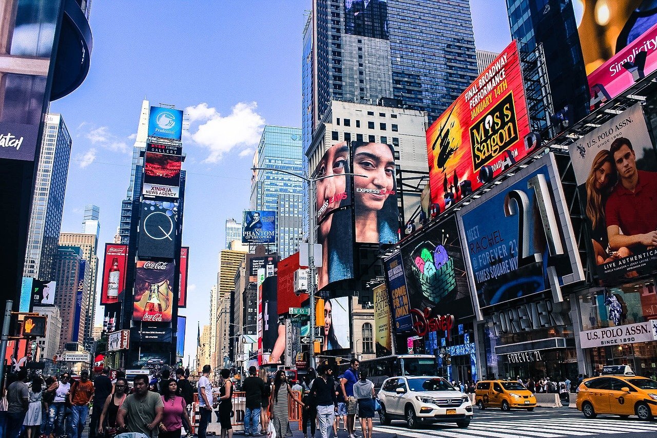 Zdjęcie przedstawia Times Square w Nowym Jorku. Po ulicy jadą liczne samochody i taksówki. Chodnikami idą tłumy ludzi. Na budynkach i wieżowcach stojących przy ulicy znajdują się liczne, rozwieszone ekrany. Widać na nich reklamy musicali filmów, Coca‑coli, ubrań.