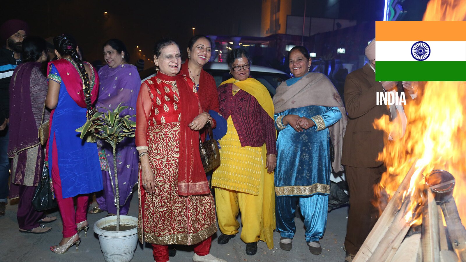 Zdjęcie przedstawia grupę stojących kobiet, ubranych w kolorowe stroje hinduskie, zwane sari, które składają się z długiej tuniki, szerokich spodni oraz szala. Pośrodku widoczne są cztery kobiety zwrócone przodem, a każda z nich ma strój w innym kolorze. Od lewej dwie w sari w kolorze czerwonym, jedna w sari w kolorze żółtym, na który ma nałożony ciemnoczerwony sweter, jedna w sari w kolorze niebieskim. Na lewo od czterech kobiet widać grupkę rozmawiających kobiet, odwróconych tyłem lub stojących bokiem. Po prawej widoczny fragment stojącego mężczyzny oraz palące się ognisko. W prawym górnym rogu widoczna flaga Indii.