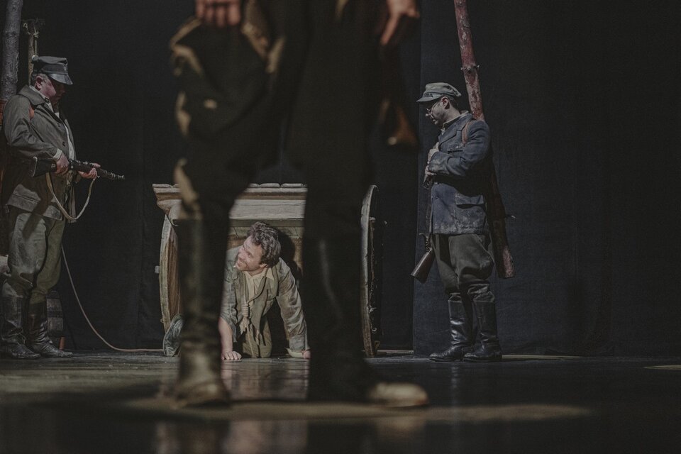 Zdjęcie ze spektaklu "Do piachu" utrzymane jest w ciemnych kolorach. Dominuje brąz. Na pierwszym planie widzimy nogi stojącego żołnierza. W tle, pomiędzy jego nogami widoczny jest mężczyzna, który uśmiechnięty wychodzi na czworakach z czegoś, co przypomina budę. Po lewej i prawej stronie wychodzącego mężczyzny stoją żołnierze. Ten po lewej trzyma w rękach karabin, lecz nie celuje do mężczyzny. Ten po prawej przygląda mu się. Lewą rękę położył na sercu, w prawej opuszczonej ręce trzyma karabin.