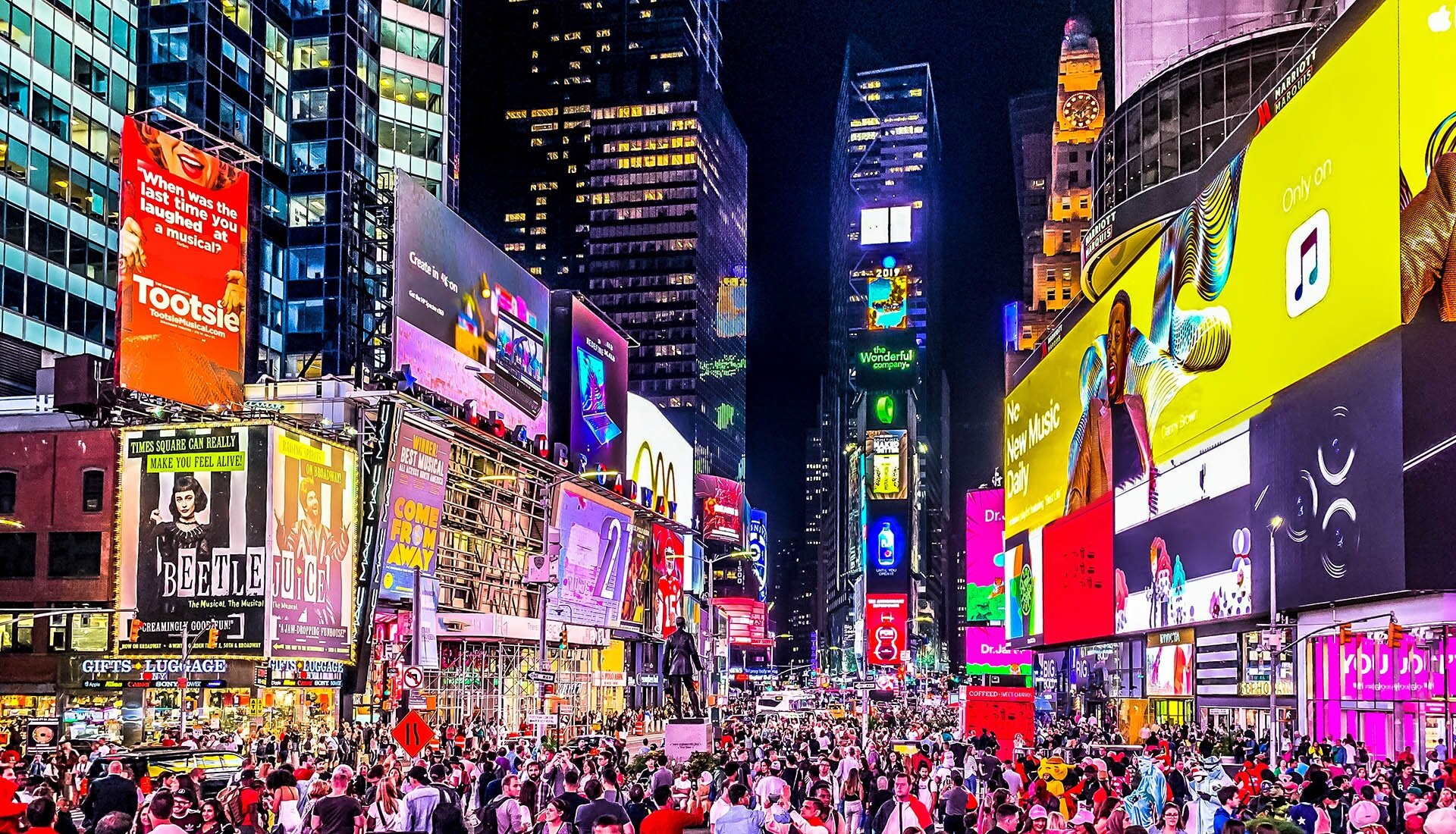 Zdjęcie przedstawia zatłoczone centrum Nowego Jorku, Times Square. Plac otoczony wieżowcami, na których wisi wiele bilbordów i ekranów reklamowych.