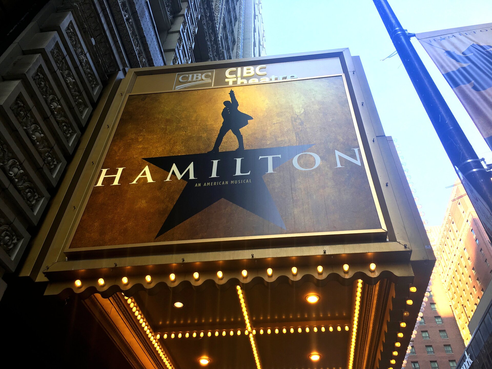 Zdjęcie przedstawia podświetloną reklamę wydarzenia zawieszoną na budynku. Na reklamie na pozłacanym tle czarna pięcioramienna gwiazda, której górne ramię to człowiek stojący w rozkroku z uniesioną do góry ręką. Poniżej napis: Hamilton an American Musical. Na górze reklamy napis: CIBC Theatre.