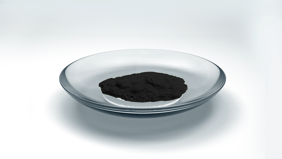 Zdjęcie przedstawia bromek miedzi(dwa) w postaci czarnego drobnego proszku umieszczonego na szkiełku zegarkowym.