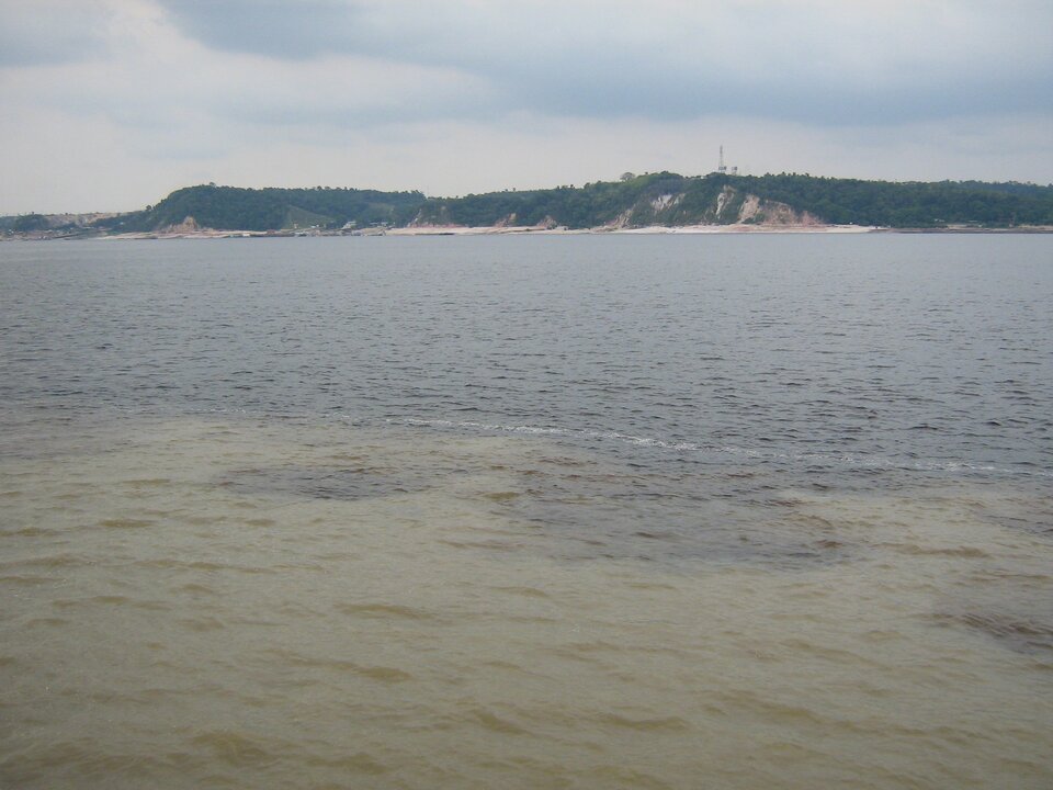 Zdjęcie szerokiego akwenu z widokiem na wybrzeże. Zdjęcie przedstawia rozległą powierzchnię wody, z linią brzegową w tle. Woda z przodu jaśniejsza, a dalej ciemniejsza. Z prawej strony widać brzeg z niskimi, zalesionymi wzgórzami.