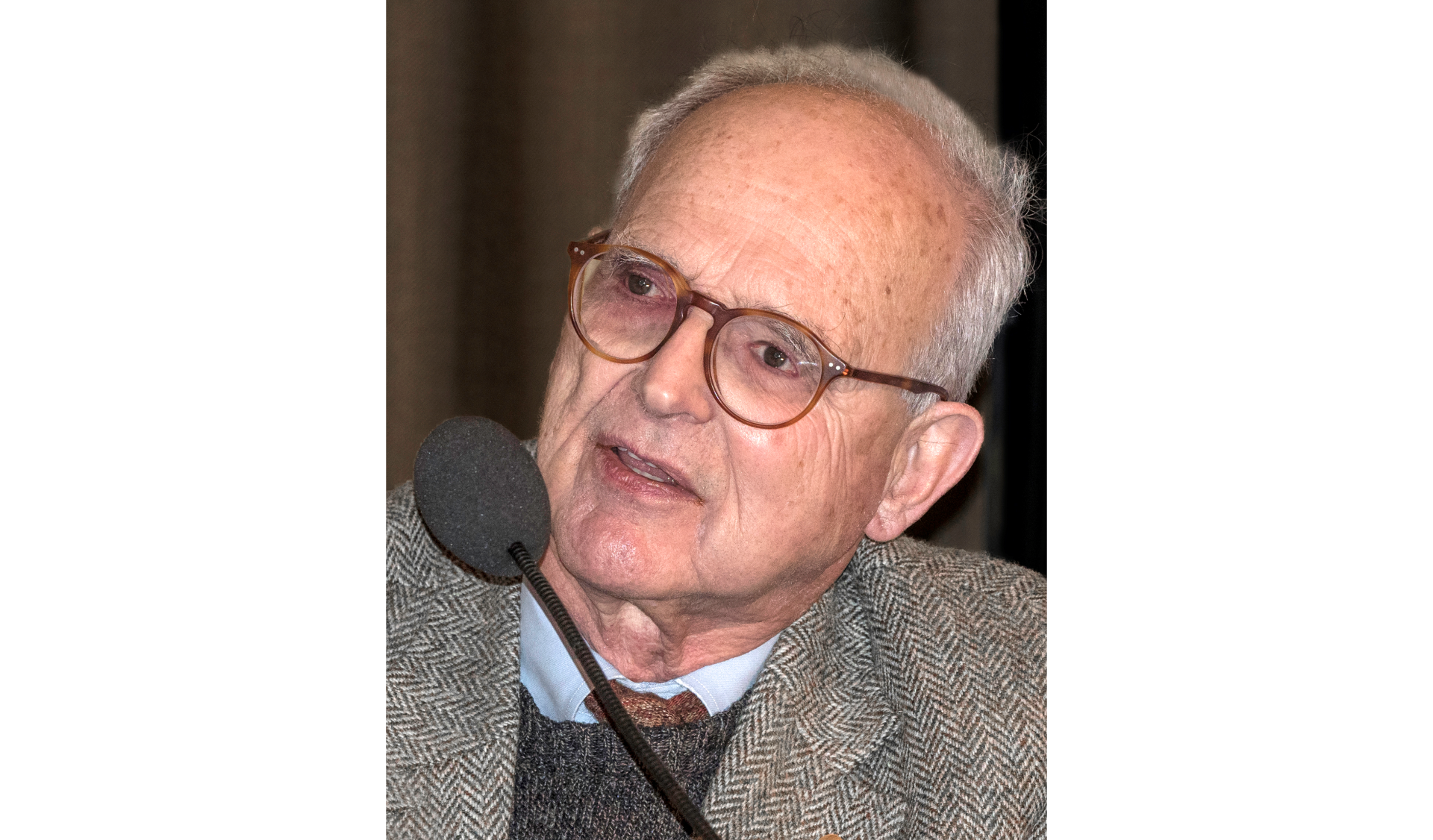 Zdjęcie przedstawia starszego pana o krótkich siwych włosach, wysokim czole, gładko ogolonego. Wysoko na nosie okrągłe okulary o brązowej oprawce. Drobne usta otwarte. Przed twarzą mikrofon.