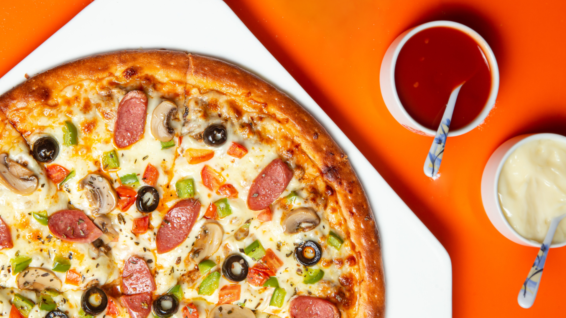 Zdjęcie wykonane z góry przedstawia pizzę po polsku. Na pizzy znajdują się: kiełbasa, papryka, oliwki, ser. Obok pizzy stoją dwa pojemniki z sosami: pomidorowym i białym.