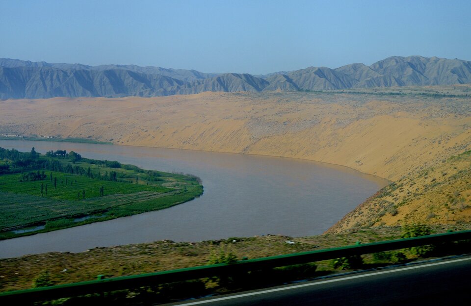 Na zdjęciu jest spokojna rzeka płynąca przez szeroką dolinę. Prawy brzeg łagodnie się wznosi, wzniesienie ma kolor żółty. W tle jest pasmo górskie.  