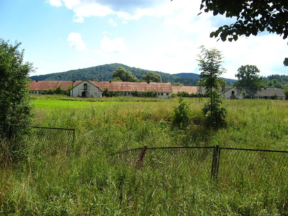 Zdjęcie przedstawia widok na PGR. Na pierwszym planie znajduje się przewrócone druciane ogrodzenie porośnięte naokoło trawą. W tle jest długi i niski budynek. Pomalowany jest na biało. Dach ma czerwony. Za nim znajduje się wzniesienie.