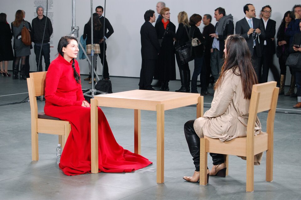 Sztuka nowoczesna Mariny Abramovic „Artystka obecna”. Zdjęcie przedstawia dwie kobiety siedzące na krzesłach przy kwadratowym stole. Zarówno krzesła, jak i stół są wykonane z jasnego drewna. Kobieta siedząca po lewej stronie ubrana jest w długą, czerwoną suknię. Ma ciemne długie włosy upięte w warkocz. Kobieta siedząca po prawej stronie ubrana jest w czarne skórzane spodnie i jasną kurtkę. Ma długie, ciemne, rozpuszczone włosy. Kobiety siedzą w przestronnym wnętrzu, w którym przy ścianach zgromadzeni są widzowie.