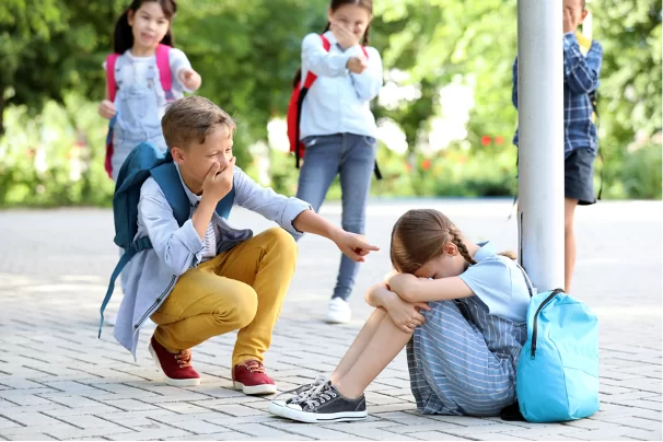 Na chodniku siedzi dziewczynka z opuszczoną głową, rękami obejmuje kolana. Niedaleko stoi troje rówieśników, wskazują na nią palcami i śmieją się. Przed dziewczynką przykucnął chłopiec i wskazuje na nią palcem, drugą dłonią zasłania usta.