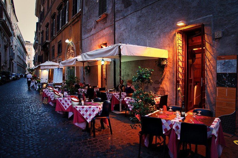 Zdjęcie przedstawia wąską uliczkę z wysokimi budynkami najprawdopodobniej mieszkalnymi. Budynki są stylowymi kamienicami. Zapada zmrok i palą się światła nad wejściami do budynków. W uliczce stoi restauracja. Stoliki nakryte są czerwonymi, białymi i czerwono‑białymi obrusami oraz sztućcami i kieliszkami. Nad stolikami są białe parasole ogrodowe. Przy jednym ze stolików siedzą zwróceni ku sobie kobieta i mężczyzna.