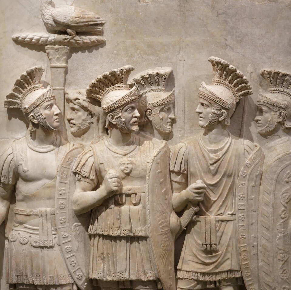 Ilustracja przedstawia płaskorzeźbę. Prezentuje grupę kilku rzymskich żołnierzy z tarczami i hełmach.