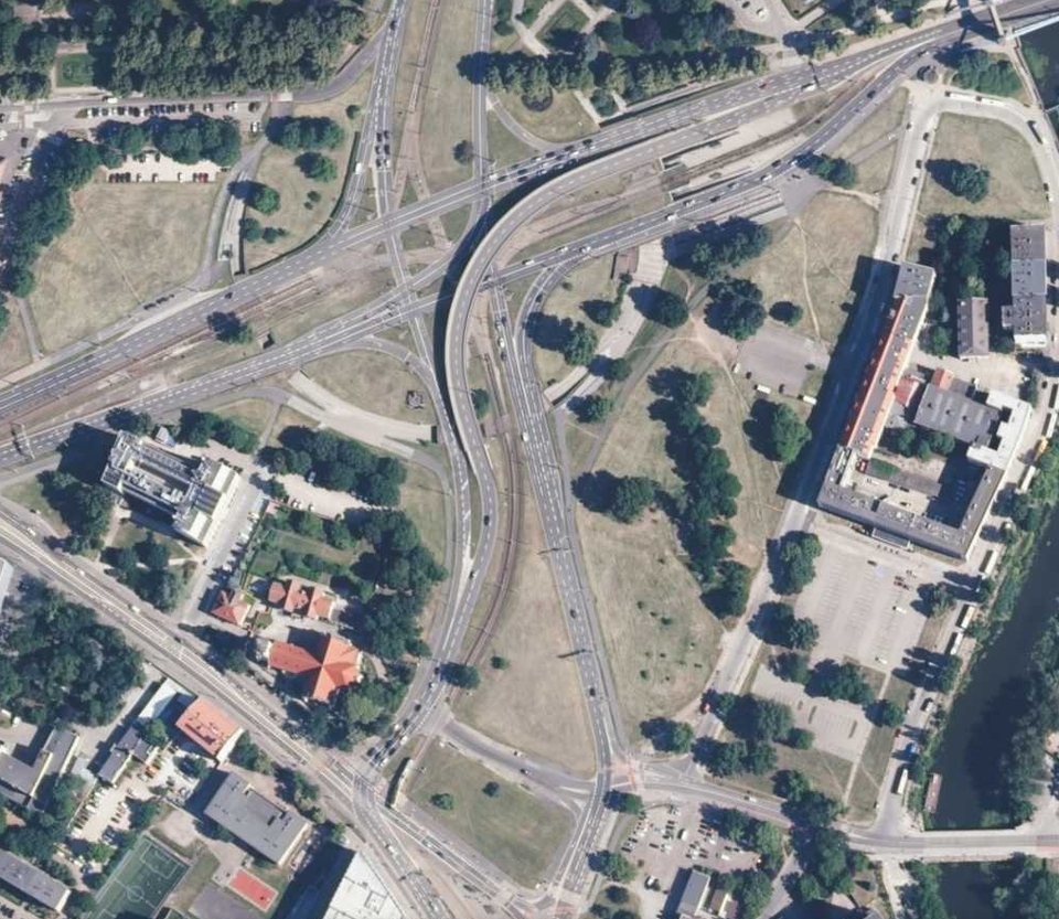 Ilustracja przedstawia zdjęcie z lotu ptaka na Plac Powstańców Warszawy we Wrocławiu. Widoczne są wielopoziomowe ulice, autostrady. Duża ilość zieleni wpasowuje się w krajobraz, widoczne są budynki mieszkalne.