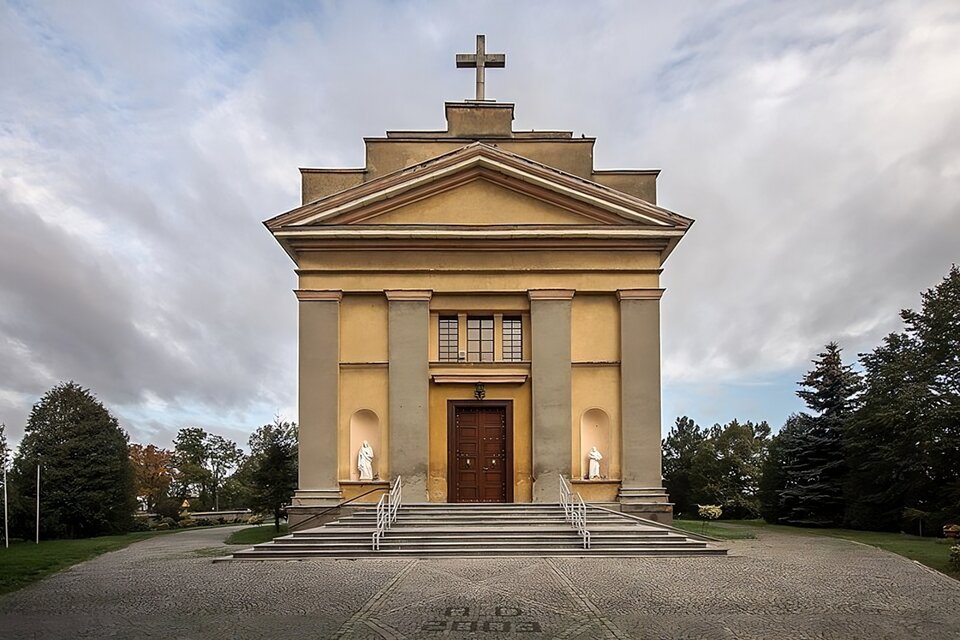 Zdjęcie przedstawia fasadę kościoła w Goliszewie. Znajdują się w niej cztery pilastry, między którymi jest portal. Całość jest zwieńczona trójkątnym frontonem i schodkową attyką. Po obu stronach wejścia są nisze z posagami świętych. Budynek znajduje się na brukowanym placu, do wejścia prowadzą niskie schody z barierkami. Wokół placu trawa, na której rosną różne gatunki drzew.