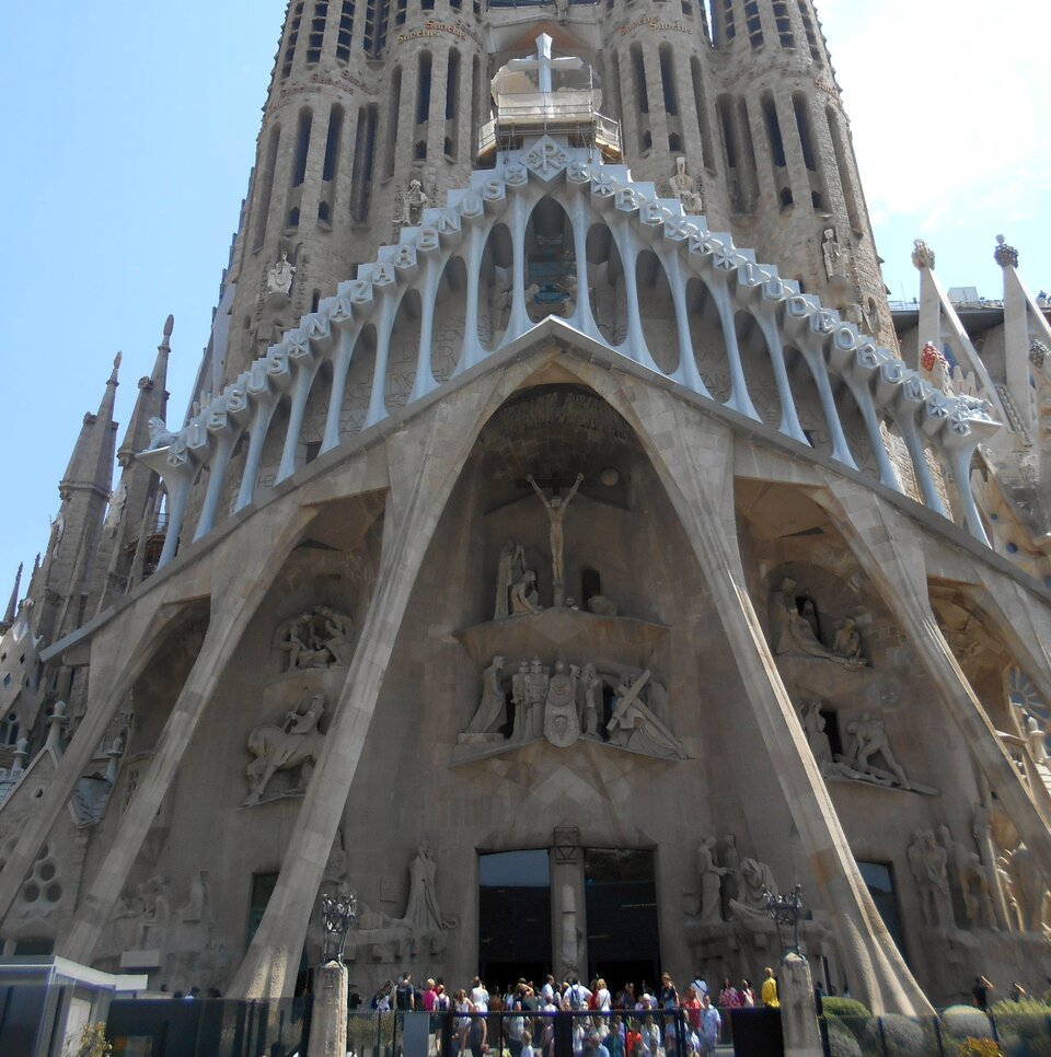 Ilustracja przedstawia Fasadę Męki Pańskiej kościoła Sagrada Família. Ukazuje surową, geometryczną fasadę o ostrych liniach i mocnych załamaniach, które podkreślają dramatyczny charakter przedstawionych scen. W centrum widoczne są rzeźby ukazujące momenty Męki Chrystusa, wyrzeźbione w kamieniu. W górnej części znajduje się scena Ukrzyżowania. Postacie mają uproszczone, kanciaste formy, co nadaje całości chłodny, ascetyczny wyraz. Poniżej widać wejście do bazyliki, a przed nim grupę zwiedzających.