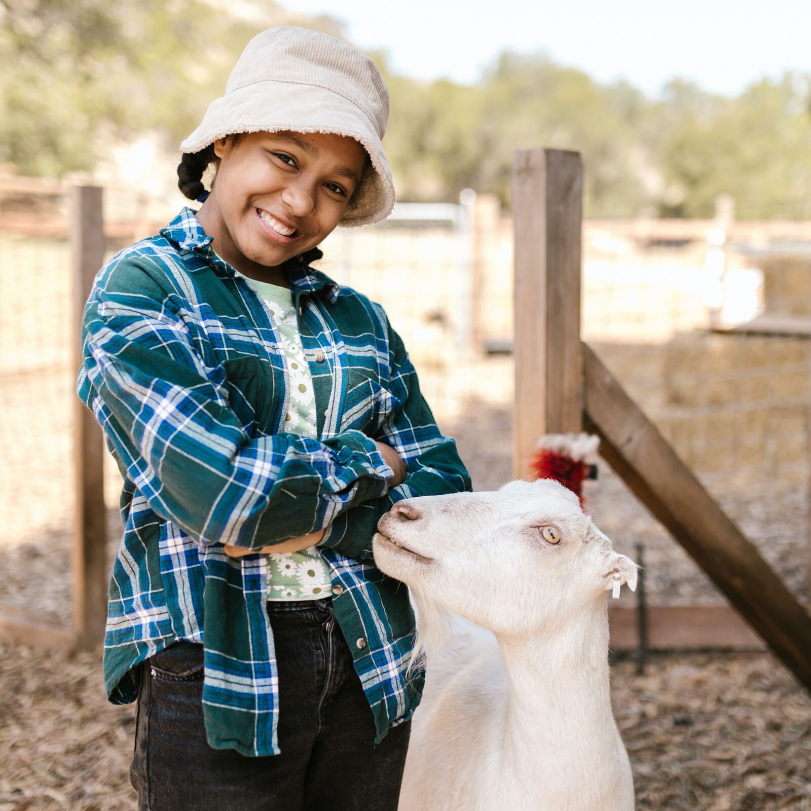Zdjęcie przedstawia dziewczynę w wieku około 11 lat stojącą w zagrodzie, a na prawo od niej stoi koza. Dziewczynka ma ciemne włosy splecione w warkocz i białą czapkę na głowie, jest ubrana w bluzkę w kwiatki, rozpiętą kraciastą koszulę i dżinsy. Dziewczynka szeroko uśmiecha się, ma głowę lekko pochyloną w prawo, a obie ręce splecione na brzuchu. Stojąca obok koza jest biała i ma głowę zwróconą w lewo i podniesioną w kierunku dziewczynki. W tle widoczne jest drewniane ogrodzenie zagrody.