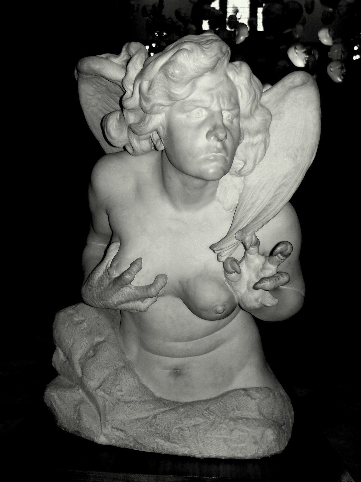 Ilustracja przedstawia fotografię rzeźby „Harpia Celeno”. Rzeźba wykonana jest z jasnego kamienia. Przedstawia popiersie półnagiej postaci, która ma głowę i ciało ludzkie oraz skrzydła i pazury ptaka. Harpia ma kręcone włosy oraz zmarszczki między brwiami. Jej lewa dłoń skierowana jest do przodu, a prawą dłonią trzyma swoją prawą pierś. Rzeźba ustawiona jest na czarnym tle. 