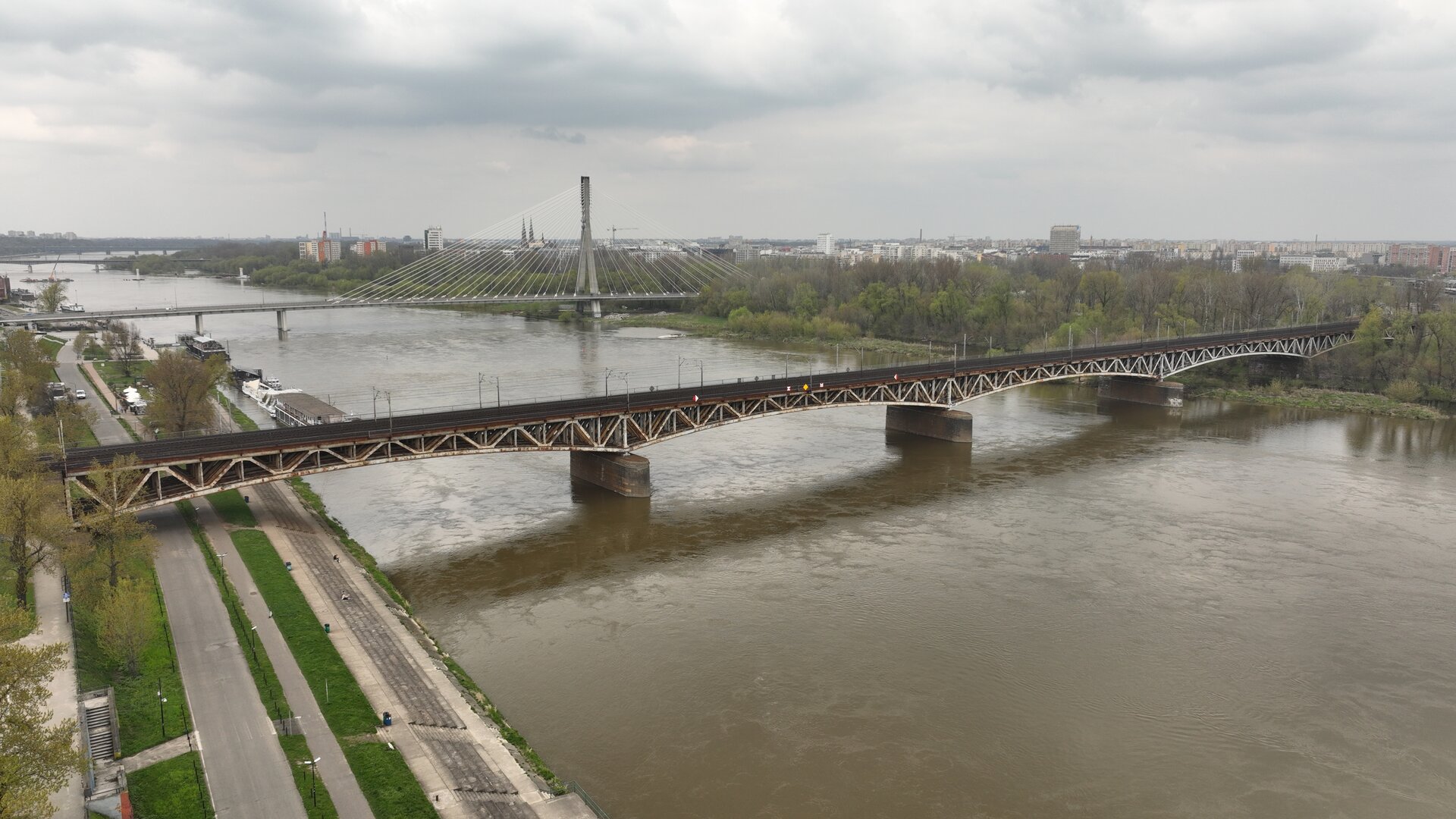  Zdjęcie przedstawia most stalowy łukowy z jazdą u góry, który biegnie nad rzeką obok dużego miasta. Konstrukcja mostu składa się z solidnych stalowych łuków, umieszczonych pod platformą, na której przebiegają tory kolejowe, a także znajdują się zabezpieczenia w postaci barierek. Most rozciąga się wzdłuż całej szerokości rzeki. Opiera się na trzech betonowych filarach umieszczonych w wodzie. W tle widoczny jest drugi most — stalowy łukowy, podwieszony, przeznaczony dla pieszych oraz samochodów. Oprócz tego na fotografii można dostrzec bulwary, wieżowce oraz inne budynki miejskie.