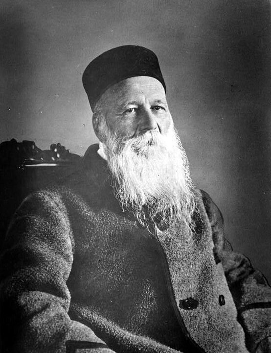 Zdjęcie przedstawia starszego mężczyznę siedzącego na wysokim krześle. Ma krótkie włosy schowane pod czapką przypominająca toczek, wysokie czoło, małe oczy, duży nos i długie siwe wąsy i brodę. Ubrany jest w dwurzędową kurtkę z grubego materiału.  