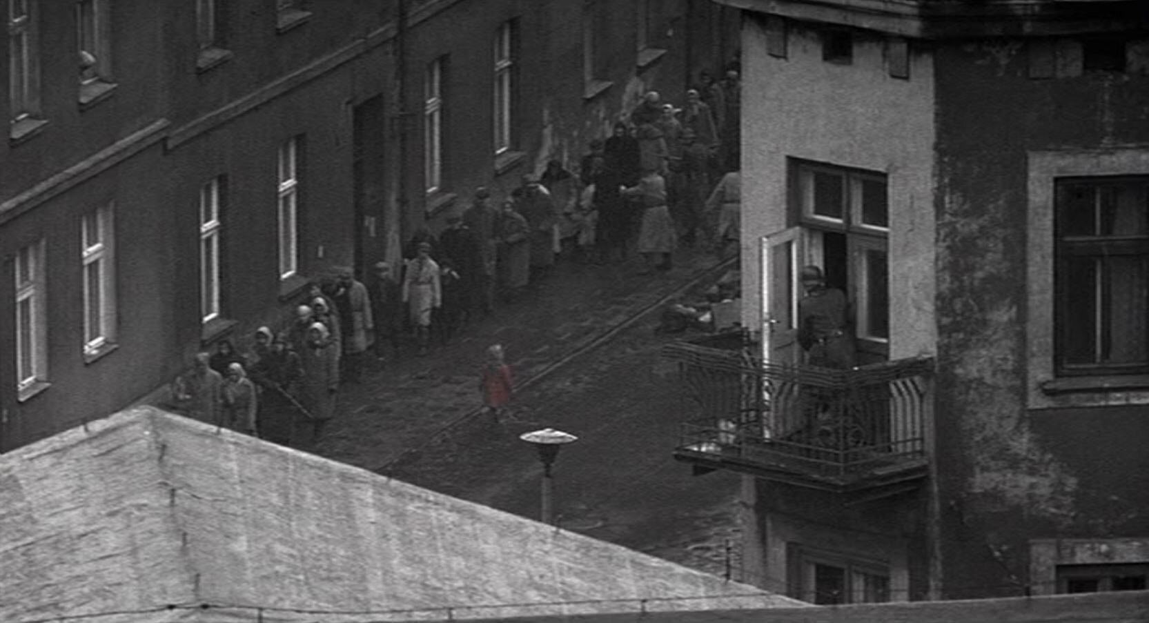 Zdjęcie czarno-białe przedstawia ulicę. Idą nią chodnikiem ludzie eskortowani przez niemieckich żołnierzy. Środkiem ulicy idzie mała dziewczynka w czerwonym płaszczyku.