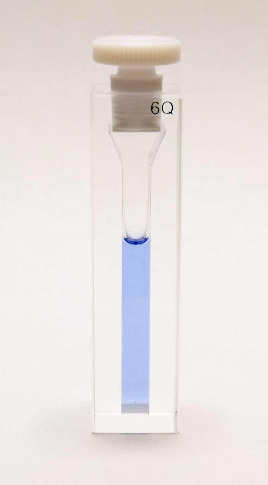 Zdjęcie przedstawia prostokątną fiolkę z małym zbiorniczkiem z jasnoniebieską cieczą. Fiolka jest zamknięta białym korkiem.