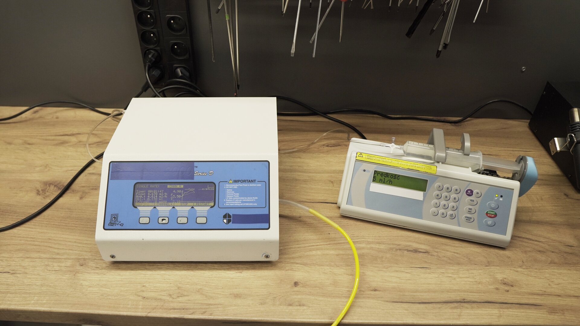 Na zdjęciu pokazane jest urządzenie testujące wraz z podłączoną pompą infuzyjną. W ramieniu pomby znajduje się strzykawka wypełniona wodą. Na ekranie pompy wyświetlana jest informacja: "Prędkość 5 ml/h".