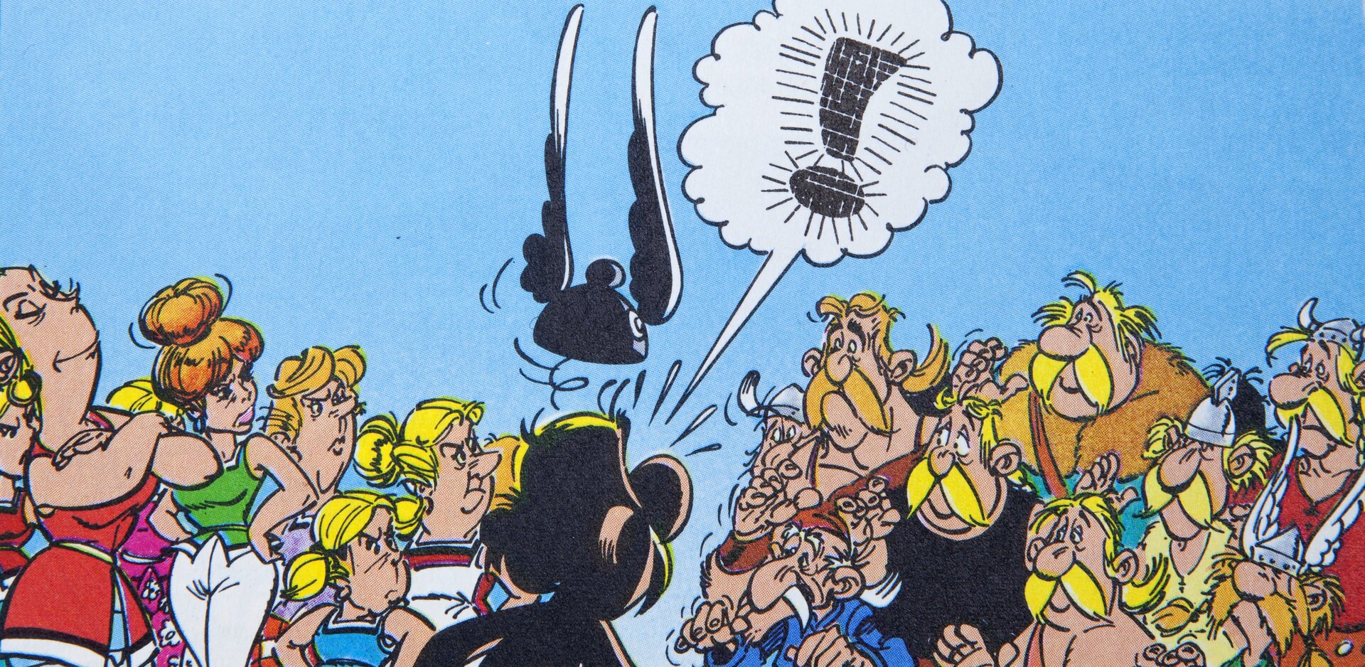 Ilustracja przedstawia fragment komiksu "Asterix. Wróżbita". Ukazuje tłum ludzi (po lewej kobiety, po prawej mężczyźni) prowadzących spór. Przed nimi stoi odwrócony tyłem do widza Asterix, nad którym unosi się jego hełm ze skrzydełkami. Nad bohaterem znajduje się dymek, w którym zamiast tekstu jest wykrzyknik.