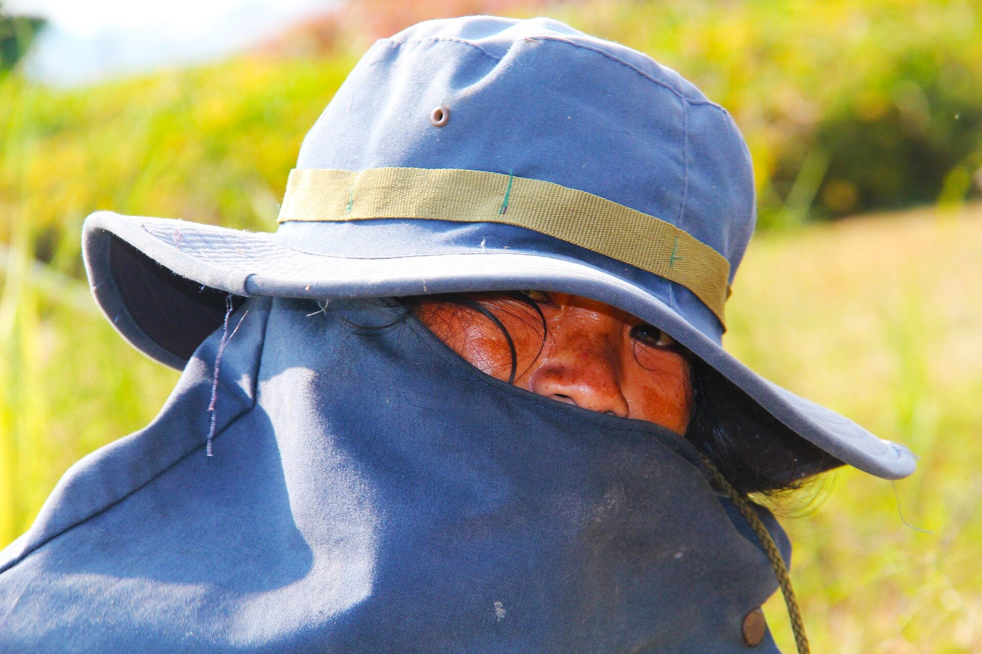 Zdjęcie przedstawia głowę kobietę, zwróconą w prawo, zerkającą w obiekty. Kobieta ma na głowie niebieski ochronnym kapeluszu z osłoną na twarz. W tle widoczne rozmyta zieleń.