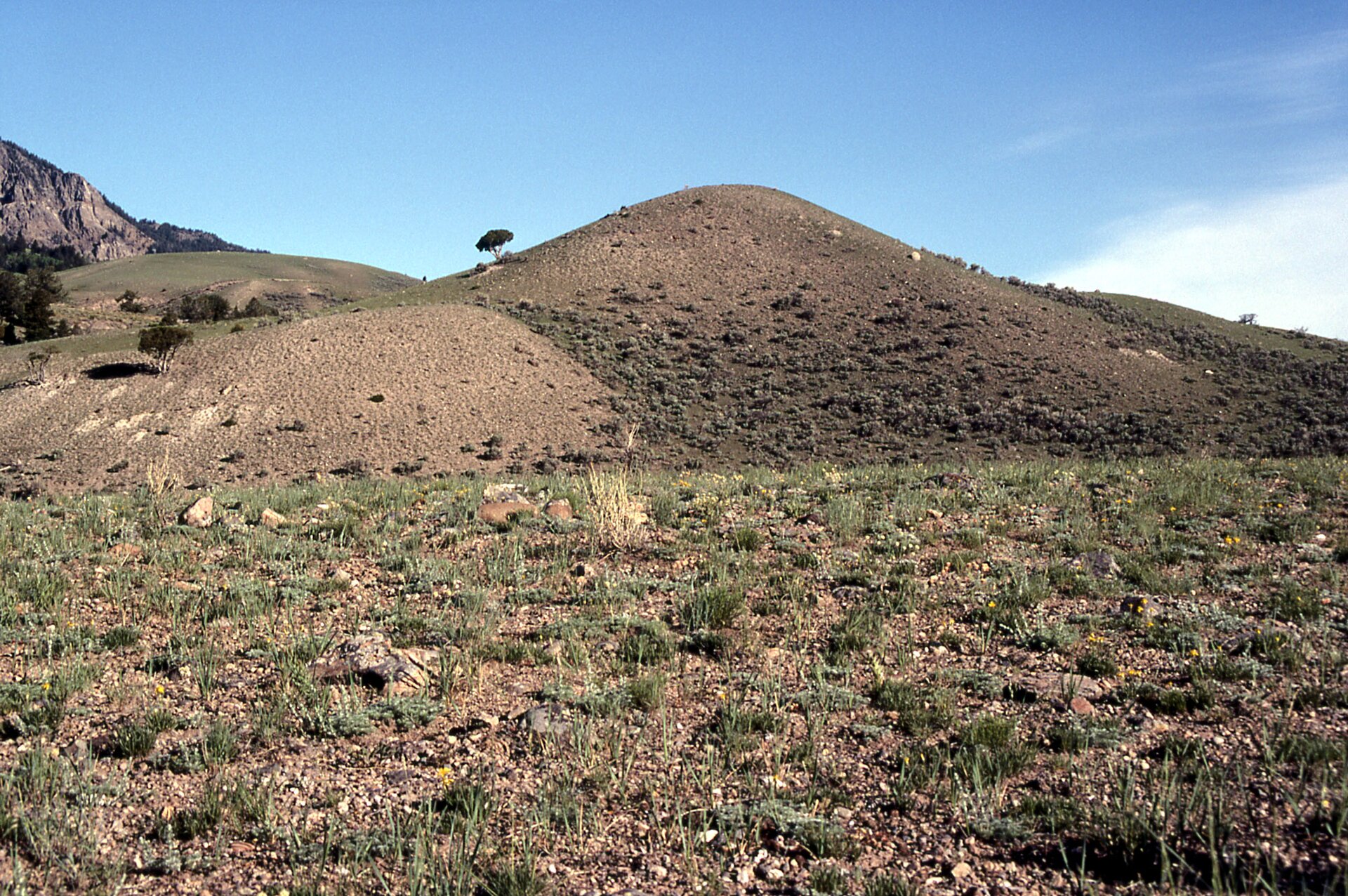 Zdjęcie B przedstawia pagórek, przypominający kształtem stożek. Ma łagodnie nachylone zbocza. Przed pagórkiem jest płaski teren, w tle są wzniesienia.  