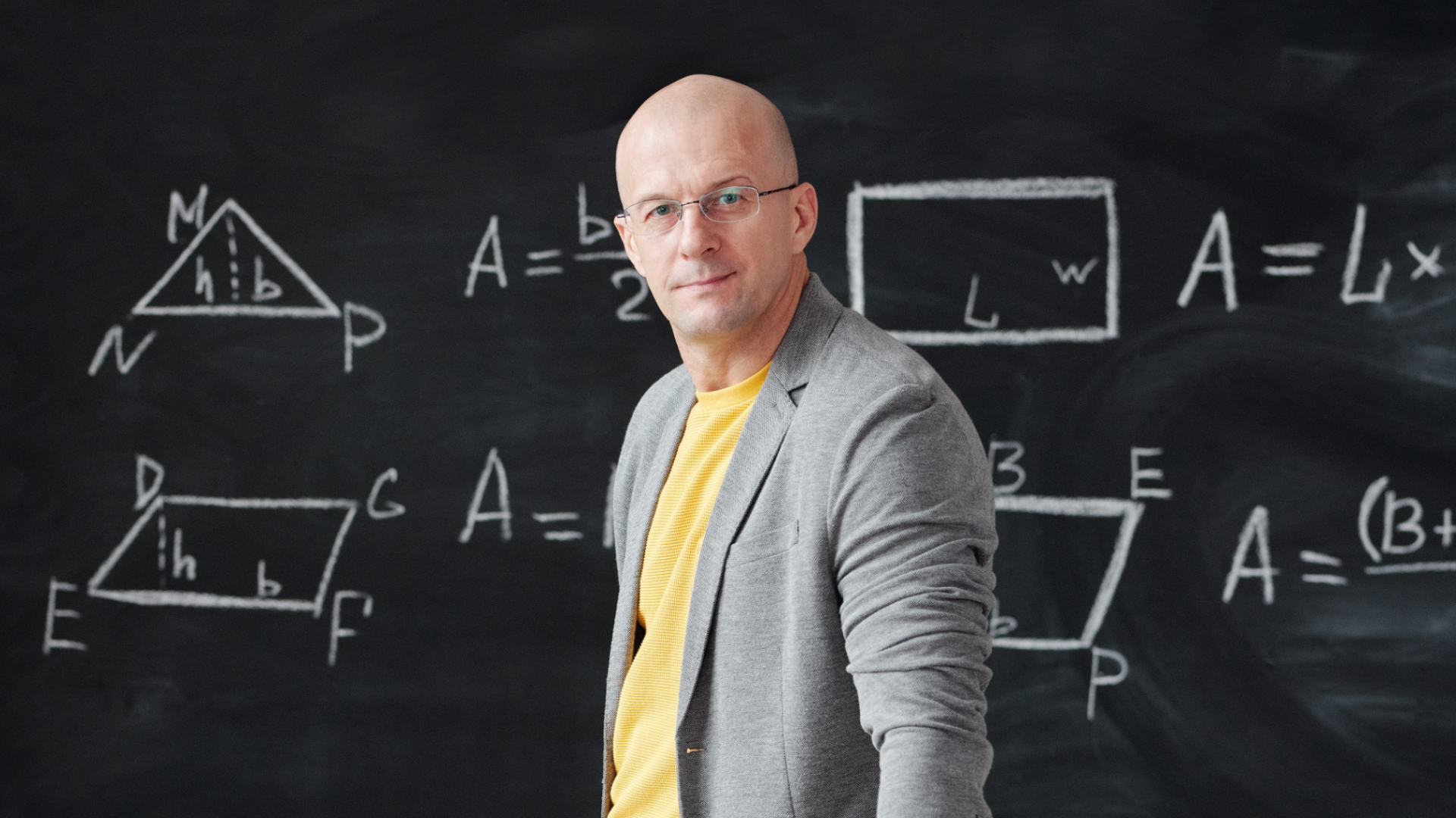 Zdjęcie przedstawia nauczyciela, który stoi na tle tablicy szkolnej. Na tablicy narysowane są figury geometryczne oraz widoczne wzory. Nauczyciel jest łysy i nosi okulary.