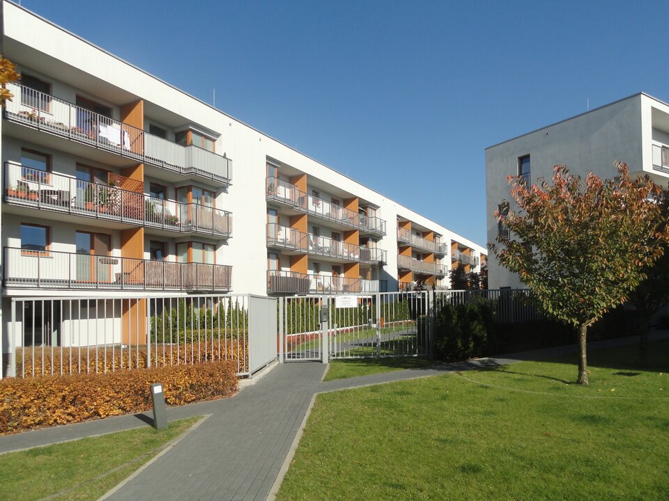 Zdjęcie przedstawia osiedle Wilno w Warszawie. Znajduje się na nim długi, biało‑brązowy blok z balkonami ze stalowych prętów. Do bloków prowadzi chodnik z kostki brukowej. Osiedle ogrodzone jest szarym, metalowym płotem. Przed ogrodzeniem na trawniku rośnie niewielkie, liściaste drzewo. 