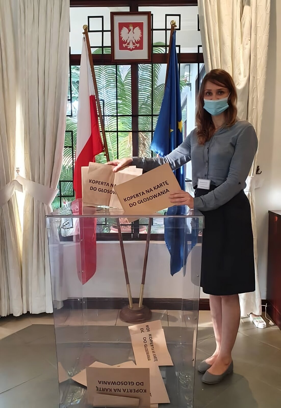 Lokal wyborczy. Widoczne godło RP.  Dwie flagi: RP i UE. Przeźroczysta urna wyborcza, w której znajdują się koperty na kartę do głosowania. Przy urnie stoi kobieta, która wrzuca koperty z kartami  do głosowania. Ma założoną maseczkę. Widoczny identyfikator.