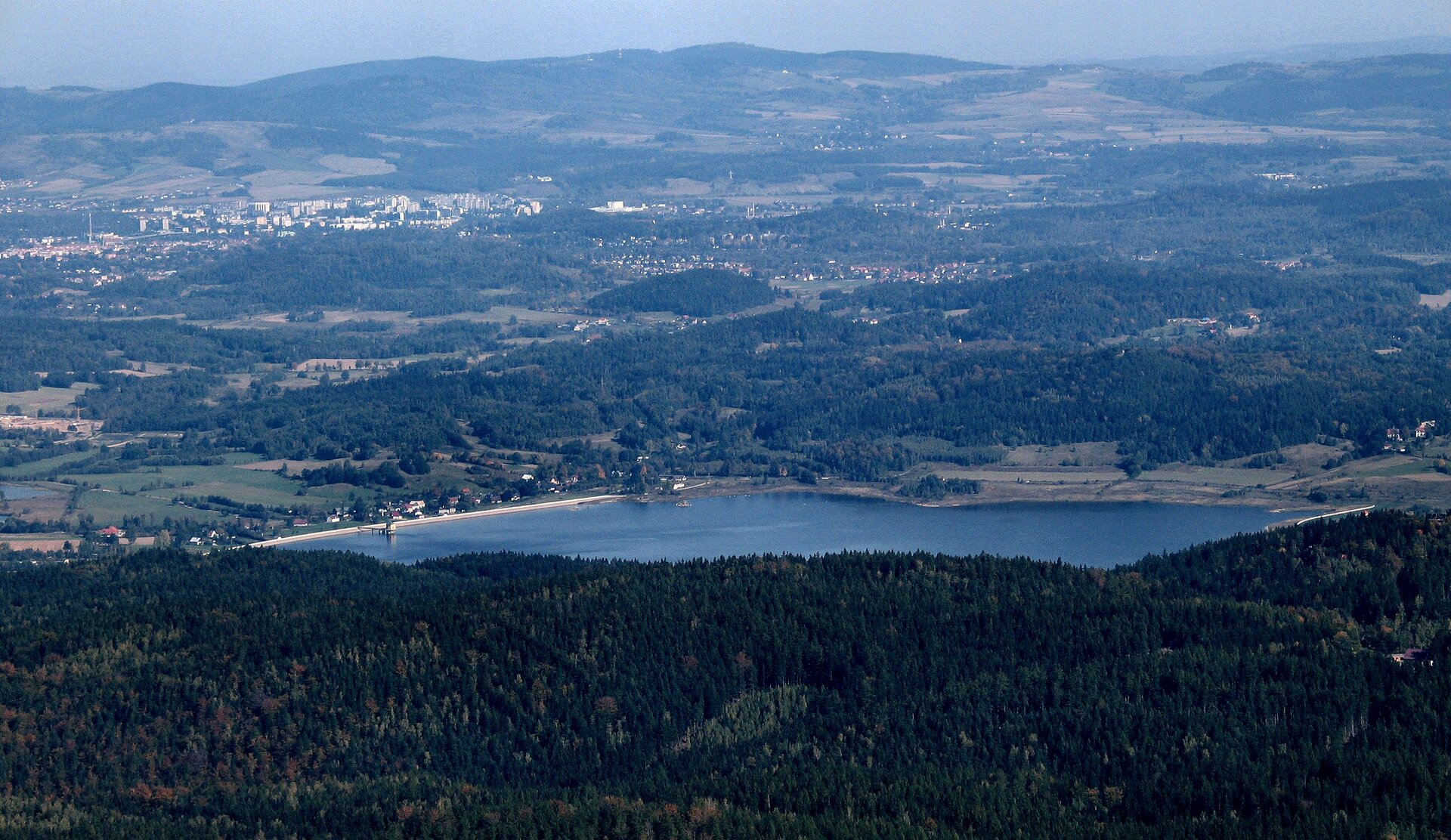 Fotografia barwna przedstawiająca pas kotlin. W środku znajduje się zbiornik wodny. Wokół jeziora widać lasy. W oddali można zauważyć zabudowania.