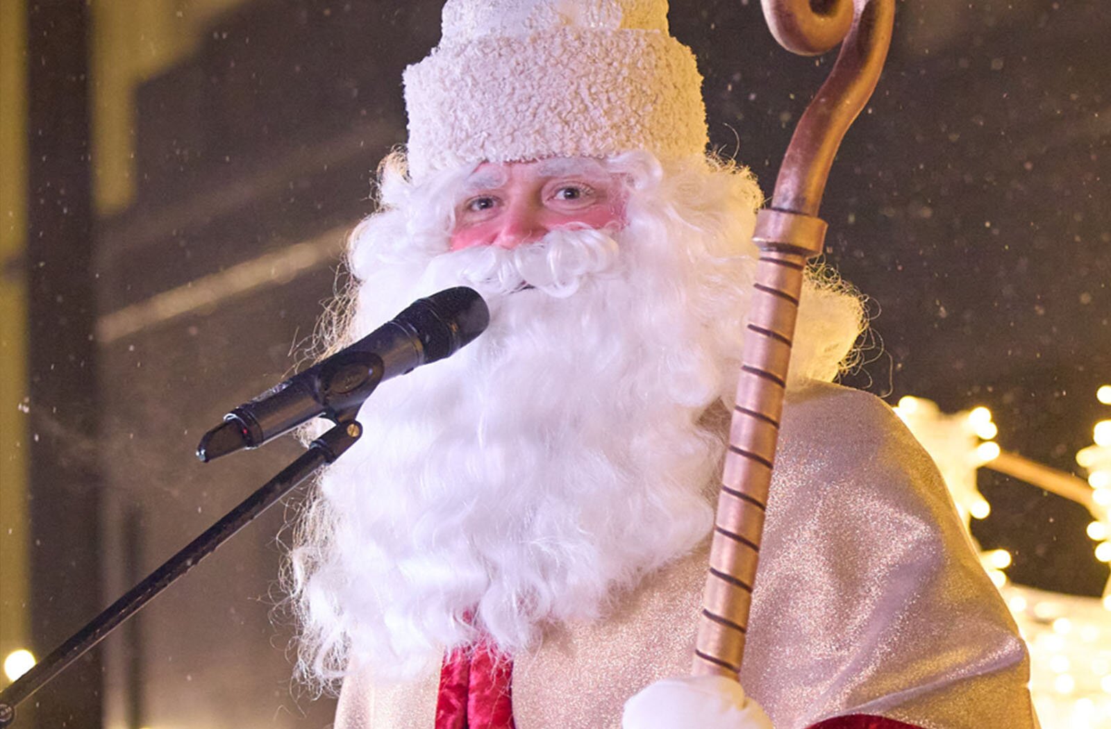 Zdjęcie przedstawia świętego Mikołaja. Widoczna jest postać mężczyzny od pasa w górę, z głową zwróconą lekko w prawo, z długimi, siwymi włosami oraz długą i siwą brodą i wąsami. Ubrany jest w złoty strój, a na głowie ma białą, futrzaną czapkę. W lewej ręce trzyma złoty pastorał.