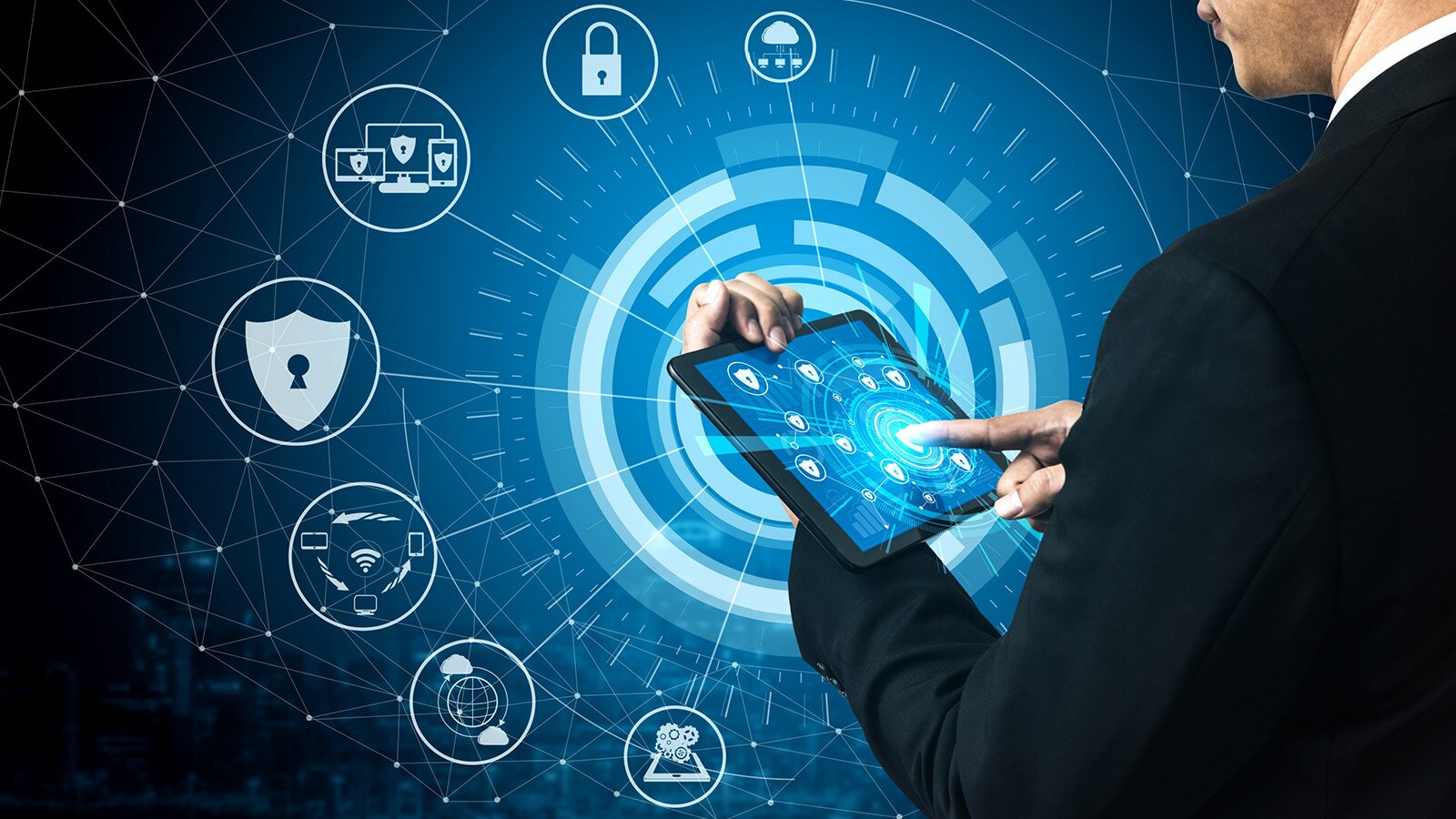 Zdjęcie przedstawia fragment postaci mężczyzny, widzianego z boku, który w lewej, ugiętej ręce trzyma tablet, zaś palcem wskazującym prawej dłoni dotyka ekranu. Na ekranie tabletu widoczne są białe ikony na niebieskim tle. W tle widoczna jest wizualizacja, która jest powiększeniem grafiki widocznej na ekranie tabletu. Wokół widoczne są różne ikony, na przykład ikona sieci internetowej, ikona tarczy z kłódką, ikona sieci WI‑Fi, ikona z tabletem, telefonem i komputerem, na wyświetlaczach których widoczne są tarcze z kłódkami, ikona zamkniętej kłódki, ikona chmury, ikona lokalizacji GPS.