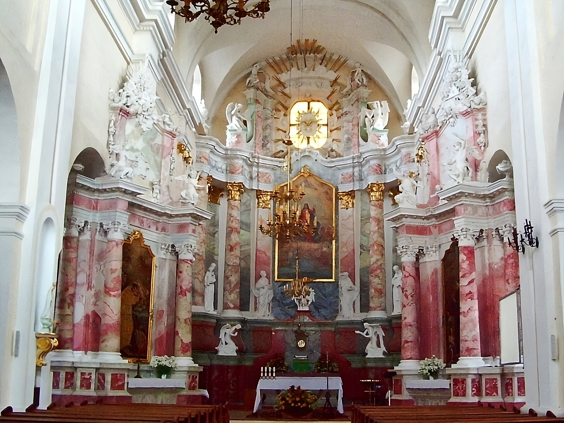 Zdjęcie przedstawia ołtarz z białego i różowego marmuru. Po prawej i lewej stronie znajdują się obrazy. Każdy z nich we wnęce, a po obydwu jego stronach kolumny. Nad nimi rzeźby. W centralnej części nieco wyżej obraz główny. Po obydwu jego stronach kolumny. Pod nim rzeźby. Nad nim okno z witraże w złotej ramie. Po jego obydwu stronach rzeźby.