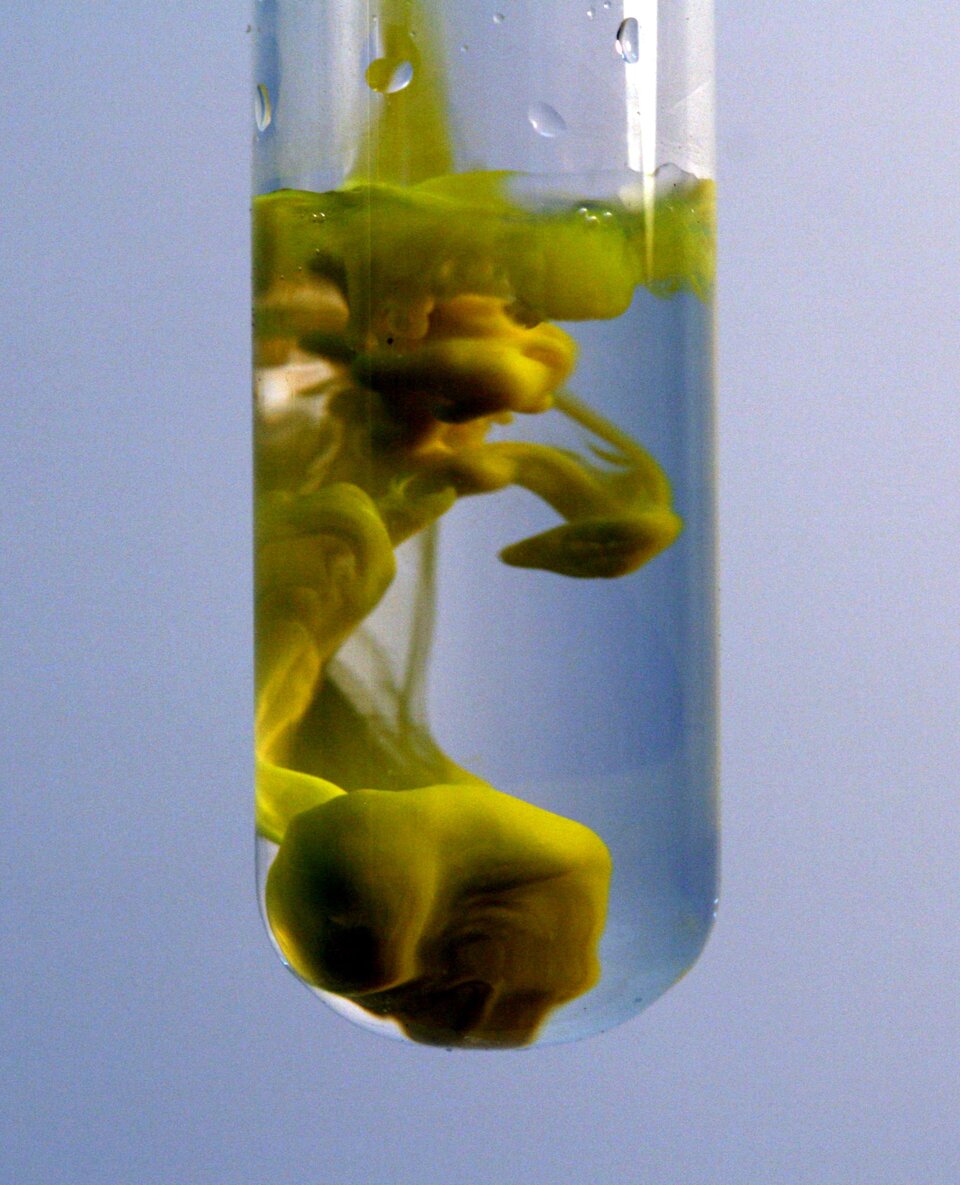 Na zdjęciu ukazano probówkę zawierającą przezroczystą ciecz. Z jej lewej strony oraz na powierzchni znajduje się intensywnie żółta, kłębiąca się niczym dym substancja.